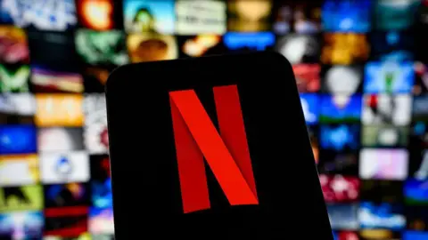 Getty Images Netflix logo shown on a smartphone screen, with the platform and its rows of film and TV shows shown blurred in the background behind it.