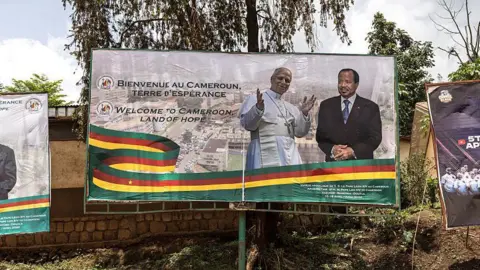 AFP via Getty Images Billboard in the centre, with slogan welcome to Cameroon, land of hope on the left and a ribbon acorss the lover half of the billboard in - green, red and yellow horizontal stripes. On the centre right is the Pope with his hands up, and next to him is the president.