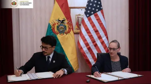 Cancillería Bolivia A man in a suit and glasses, with short dark hair, holds the arms of a woman wearing a shirt and suit jacket and glasses, in front of the American and Bolivian flags.