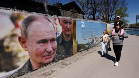 Reuters A poster showing Russian President on the side of a road where a man wearing black trousers, a gret top is carrying a child on his neck, with a woman walking by his side, wearing light blue trousers,a white top and a pink jacket, as well as carrying a small black handbag 
