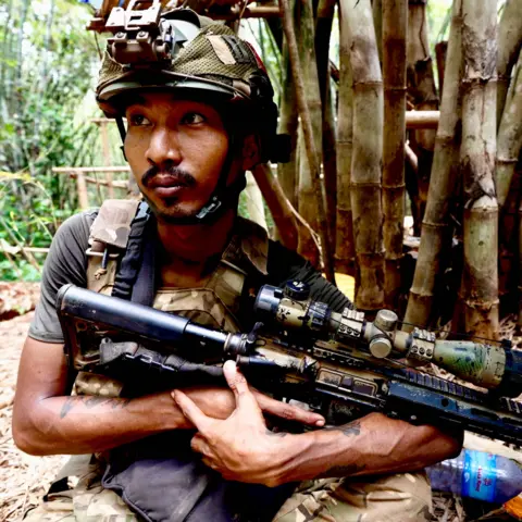 BBC/Darren Conway A soldier in a camouflage helmet holds a gun in a jungle setting, a tree trunk behind him. He has a watchful expression and his eyes are fixed beyond the camera.
