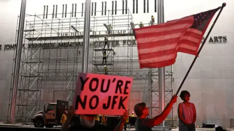 AFP via Getty Images people gathered in front of Kennedy Center building, one holding an American flag and another holding a sign that says 