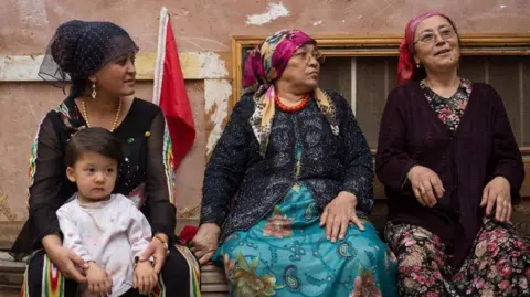 Getty Images A young Uyghur women, with a small child on her knee, sits next to two older women on a street in Kashgar City, Xinjiang. All three are dressed in traditional garments with head scarves