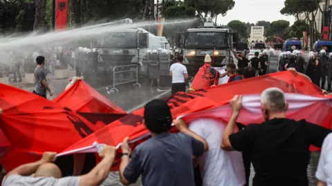 Reuters Water cannons are fired at a row of protesters holding Albanian flags from two large police vehicles in a city square.