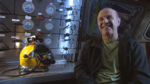 BBC Scotland Doug Allan smiling at the camera. He is sitting down and is wearing a dark, open jumper over a green t-shirt. A yellow divers helmet is on the table next to him.