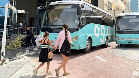EPA/Shutterstock A woman and a girl walk in front of buses prepared for stranded airline passengers outside a hotel in Doha, Qatar. Photo: 1 March 2026