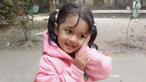 Supplied: Al Amin A young girl wearing a pink hooded jumper with her hair in pig-tails holds a hand against her face, smiling
