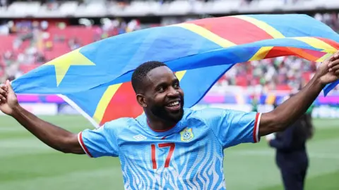 Getty Images Congolese football player Cédric Bakambu celebrates with the national flag on a football pitch