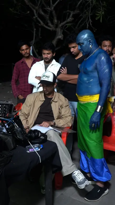 Don Vanzara Showbiz LLP A bald man wearing blue body paint, a green skirt and bright blue pants stands and previews a recorded scene with other people around