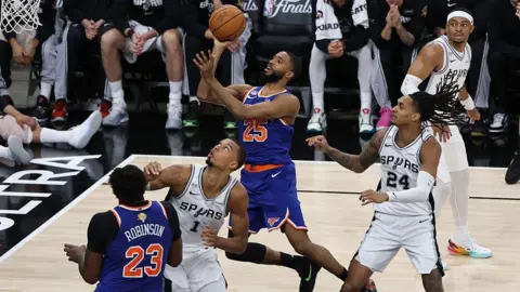 NBAE via Getty Images A Knicks player leaps towards the basket with the ball in hand as opposing Spurs players surround him