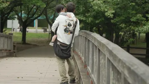 Jiro Akiba/BBC A man dressed up in a hoodie carries a baby in his arms while crossing a bridge in a park.