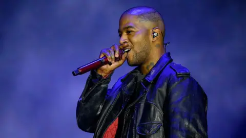 Getty Images Kid Cudi is wearing a black leather jacket, he is singing into a handheld mic looking off to the left. He has a shaved head and brown eyes.