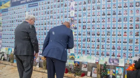 Ghana's Foreign Affairs Ministry/Facebook Ukraine Foreign Minister Andrii Sybiha‎ in black suit and his Ghanaian counterpart Samuel Okudzeto Ablakwa in blue suit bow their heads in respect before a memorial wall honouring those killed in the war, as snow falls gently around them