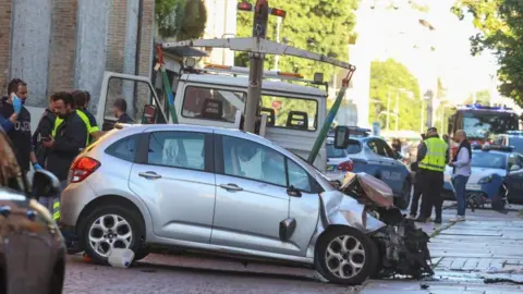 Reuters A silver car with the front mangled up next to a recovery vehicle, with police and police cars visible in the middle of the road