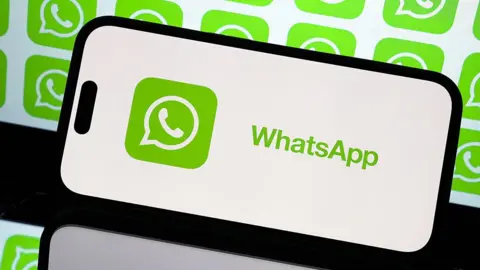 Anadolu via Getty Images WhatsApp's logo displayed against a white background on a smartphone, with a reflection of the screen appearing below. Behind the smartphone is a larger display of repeated WhatsApp's green logos, which depict a telephone inside a message bubble.