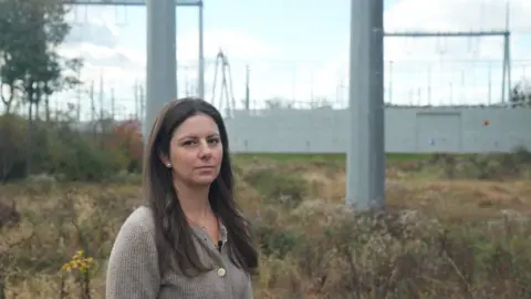 Meiying Wu Emily Kasabian stands in front of a substation with giant poles. 