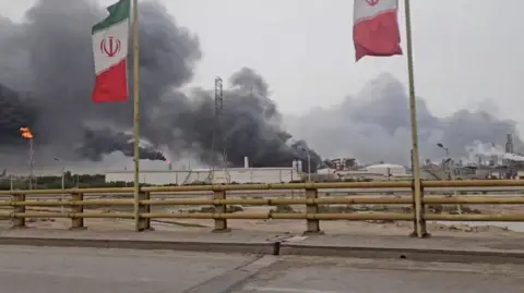 Reuters Smoke rises after strikes on the Mahshahr Petrochemical Zone in Bandar Mahshahr County, Khuzestan Province, Iran. Two flags flutter on poles in the foreground on the side of a road with the attacked plant behind 