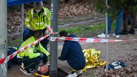 Reuters Man lying on the ground receives medical attention from three emergency workers, two of them in high-viz jackets, with police tape in the foreground