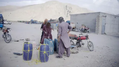 Men attaching ropes and string to large blue gallons of fuel. They're surrounded by a grey, dry landscape and a couple of motorbikes. There's a mountain in the background