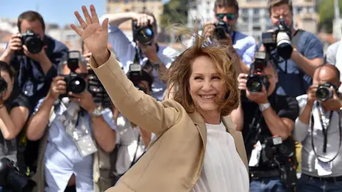 Getty Images Nathalie Baye, a woman with short brown hair, waves in front of a crowd of photographers in Cannes. She wears a camel-coloured blazer and a white t-shirt. She is smiling.