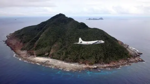 Getty Images A white plane tilts in the air as it passes a verdant hilly island part of the Senkaku/Diaoyu island chain. 