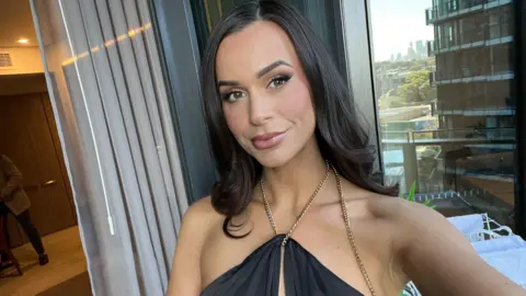Sierah Swepstone A woman with long dark hair wearing a sleeveless top. She is smiling, and next to a window that looks out on an apartment building and a sunny cityscape.