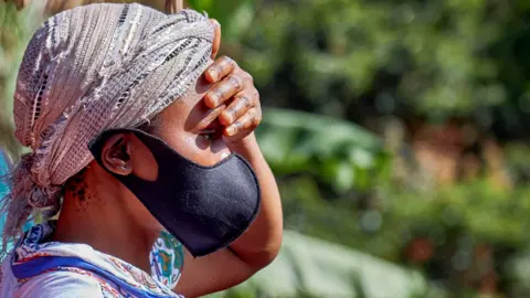Reuters A head-only side profile of a woman facing right. she has a scarf wrapped around her head and is wearing a black protective face mask. She has her left hand over her eyes to shield from the sun.