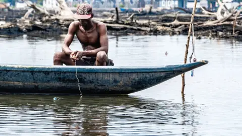 Simi Jolaoso / BBC A man sits on a boat holding a fishing line in the waters of the Niger Delta. Behind him can be seen dead mangroves and some blurry buildings.