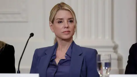 Getty Images Former US Attorney General Pam Bondi sits at a table behind a tabletop microphone as she listens to US President Donald Trump speak during a lunch with the Trump Kennedy Center Board Members on 16 March 2026.