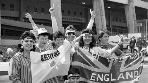 Monte Fresco/Daily Mirror/Mirrorpix via Getty Images English and Argentine fans share a laugh and a joke outside the Azteca stadium. The photo is black-and-white and they are holding banners from their respective countries and smiling broadly. 