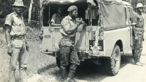 Godwin Alabi-Isama A Nigerian soldier leans against an army truck stopped by the side of a road - thick vegetation seen behind - and speaks into a walkie-talkie. Another combatant can be seen in the back of the vehicle beneath a canvas canopy and two other soldiers are seen standing nearby. It is a black and white archive shot.