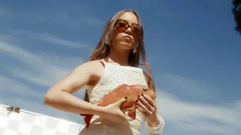 Laufey/Vingolf/Awal Laufey, framed against a sunny Californian sky, holds a red snapper while preparing to film a music video for her single Mad Woman