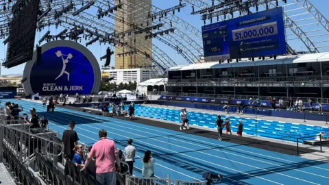 BBC Athletes aalk around a blue athletics track and 4-lane swimming pool as the golden Trump Tower looms in the background. A sign advertises a million dollars in prize money for athletes at the Enhanced Games. 