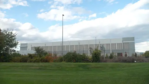 Meiying Wu A large industrial warehouse-type building sits behind a flat grassy field and trees.