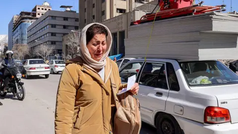 AFP via Getty Images A woman appears to be crying as she walks on a street in Tehran. She has a light coloured headscarf and a tan coat on. She is holding a piece of paper in her hand and has a rucksack on her shoulder. A motorcyclist is behind her and cars line the street to her left.