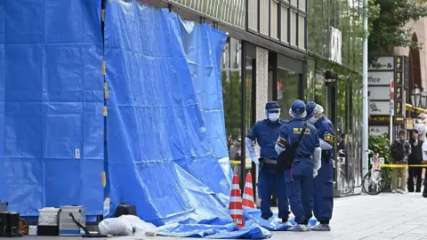 Getty Images Three people wearing a navy blue uniform and protective gear stand on a street in front of a large shopping centre. Next to them is a large blue tarp sheet with two red and white cones. Behind them is a yellow emergency cordon. 