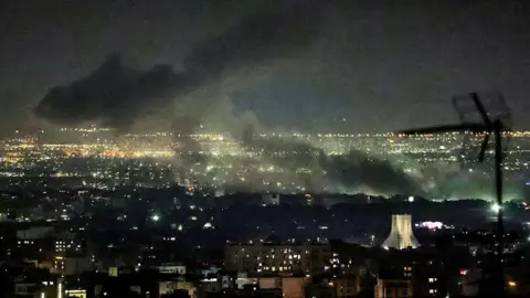 AFP via Getty Images Smoke plumes billow from the site of airstrikes near Azadi Tower in western Tehran on 10 March. The night sky and smoke are black. Below, lights are on in buildings and a tower is lit up.