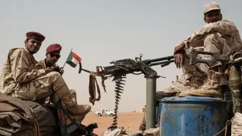 AFP via Getty Images Three soldiers sitting on machinary in a dessert terrain. Theu are wearing uniform. The two on the left have burghundy beret and one is holding a small flag. The man on the right is wearing a cap