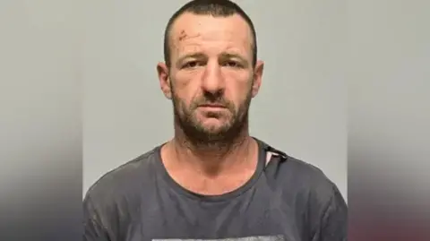 NSW Police Force NSW Police Force Headshot of suspect Julian Ingram, who is seen with a beard and wearing an old grey T-shirt.