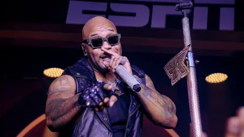 Getty Images A man wearing rhinestone-studded sunglasses sings into a rhinestone studded microphone while he points with his right hand