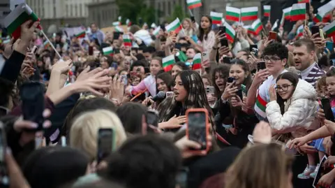 Getty Images A large number of people are gathered to welcome Dara home in Sofia. Many of them have their phones out to take photos of her and a number of people in the crowd are waving Bulgarian flags. The singer herself is visible among the crowd, talking into a microphone.