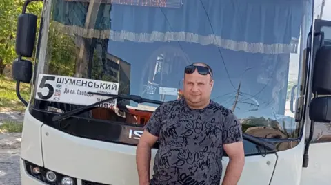 BBC A man stands in front of a bus with the number 5 on the front wearing sunglasses and a grey t-shirt.
