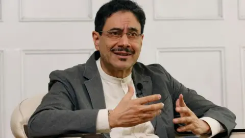 EPA Iván Cepeda gestures with his hands as he sits on a beige-upholstered chair. He is wearing a grey jacket over a collarless white shirt. He has dark hair, a moustache and is wearing glasses. Behind him is a white-panneled wall.
