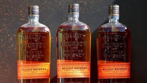 Getty Images Three bottles of Bulleit Bourbon are lined up against a black stone background The labels are orange and printed with vintage-style writing, while the bottles, filled with golden liquor, are embossed with the words "Bulleit Bourbon: Frontier Whiskey"