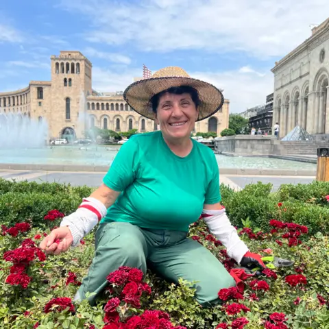A smiling woman, wearing a straw hat, green t-shirt and trousers and white arm coverings,kneels in a bed of roses with stone buildings and a fountain behind her