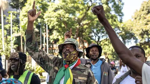 AFP via Getty Images A man in a camouflage top and South African flag around his neck holds his right hand in the air with his index finger raised. 