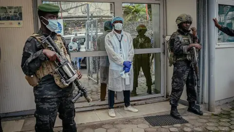 AFP via Getty Images Two armed soldiers standing in front of a lab. In front of the door is a doctor in protective clothing. 
