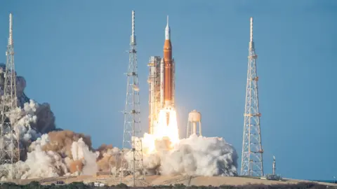NASA A tall white and orange rocket is blasting off from a coastal launch pad. Flames and bright white exhaust pour from its base, creating a wide, glowing tail that hides the ground in thick clouds of smoke. The rocket is pencil‑thin and points straight up into a clear blue sky, with the sea faintly visible behind it. Slender white booster rockets cling to either side of the central orange core. To the left and right stand two latticework metal towers, like giant scaffolding poles, framing the rocket. A rounded white tank sits nearby on spindly legs, partly lost in the steam. The overall impression is of immense power and light as the vehicle climbs away, leaving a boiling, churning cloud where it stood.