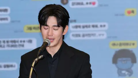 WireImage A tearful Kim Soo-hyun looking down and speaking into a microphone. He is wearing a black shirt and black blazer. Behind him is a projection of screenshots of text messages.