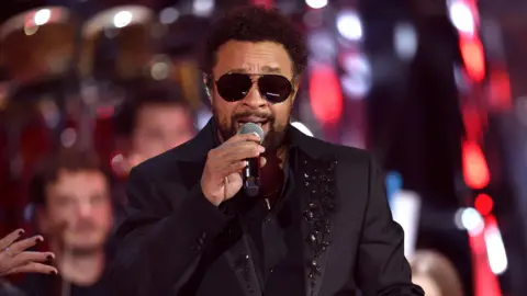 Getty Images Shaggy performing in 2021, wearing a black, bejewelled suit jacket and sunglasses.